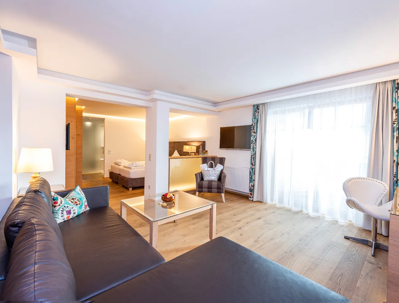 Modern hotel room with wooden floor, seating area, TV and large window with curtains